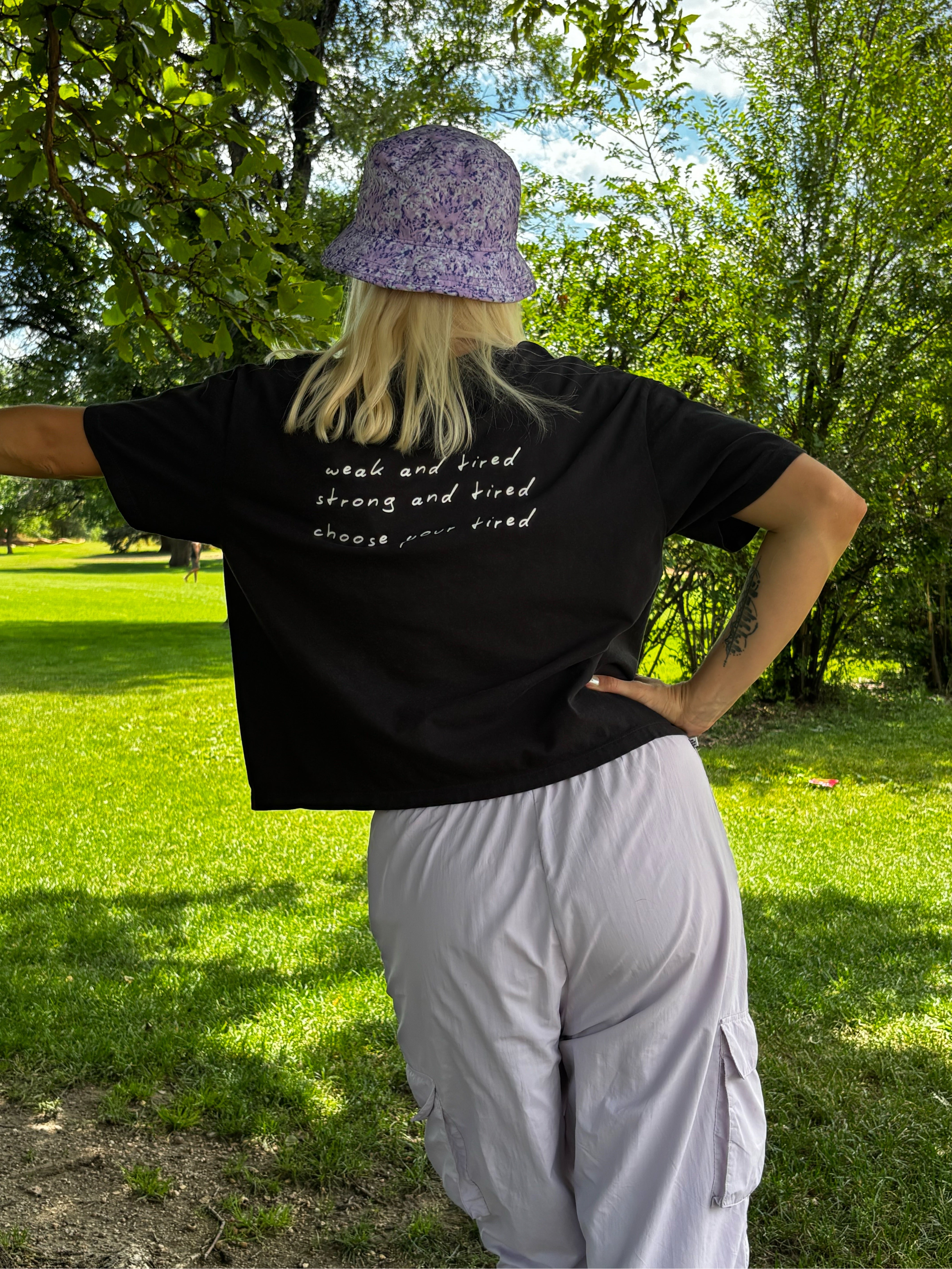 A person standing outdoors in a park, viewed from the back, wearing a black graphic T-shirt with white script text, a purple patterned bucket hat, and light gray cargo pants.