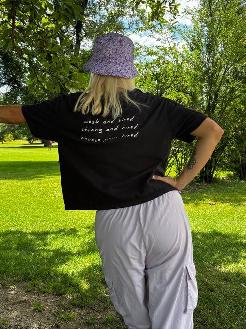 A person standing outdoors in a park, viewed from the back, wearing a black graphic T-shirt with white script text, a purple patterned bucket hat, and light gray cargo pants.